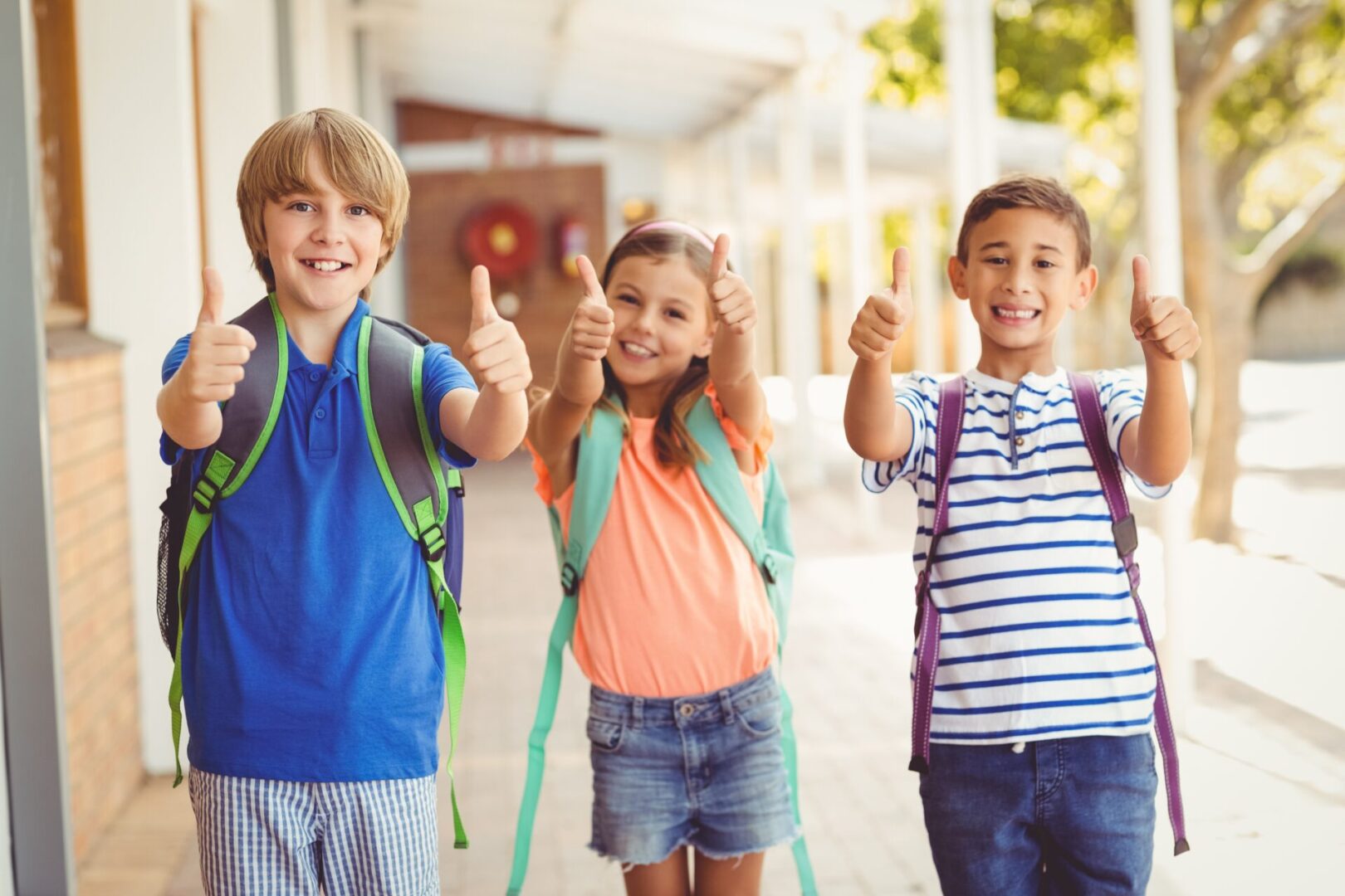 Three children giving thumbs up in front of a school building.